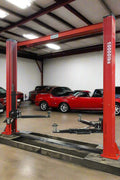 2-post vehicle lift in red installed in garage

Close-up of lifting arms and safety lock mechanism

Base of hoist with truck tires for scale

Top bar and hydraulic system – JH5000F model